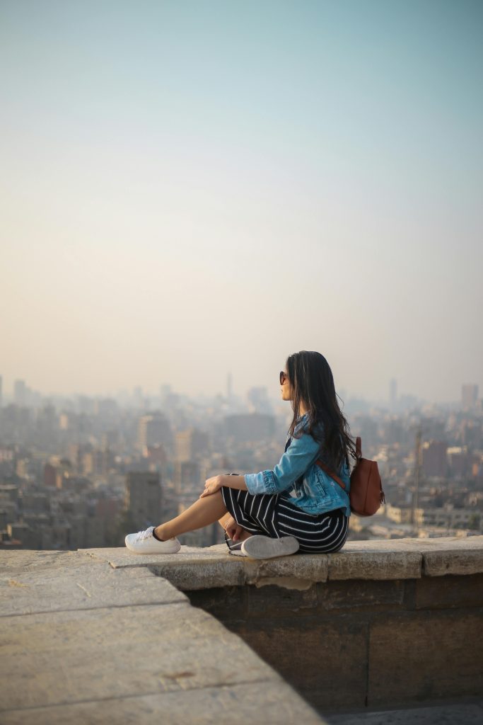 A young woman in casual attire sits on a rooftop terrace, viewing a sprawling cityscape in daylight.