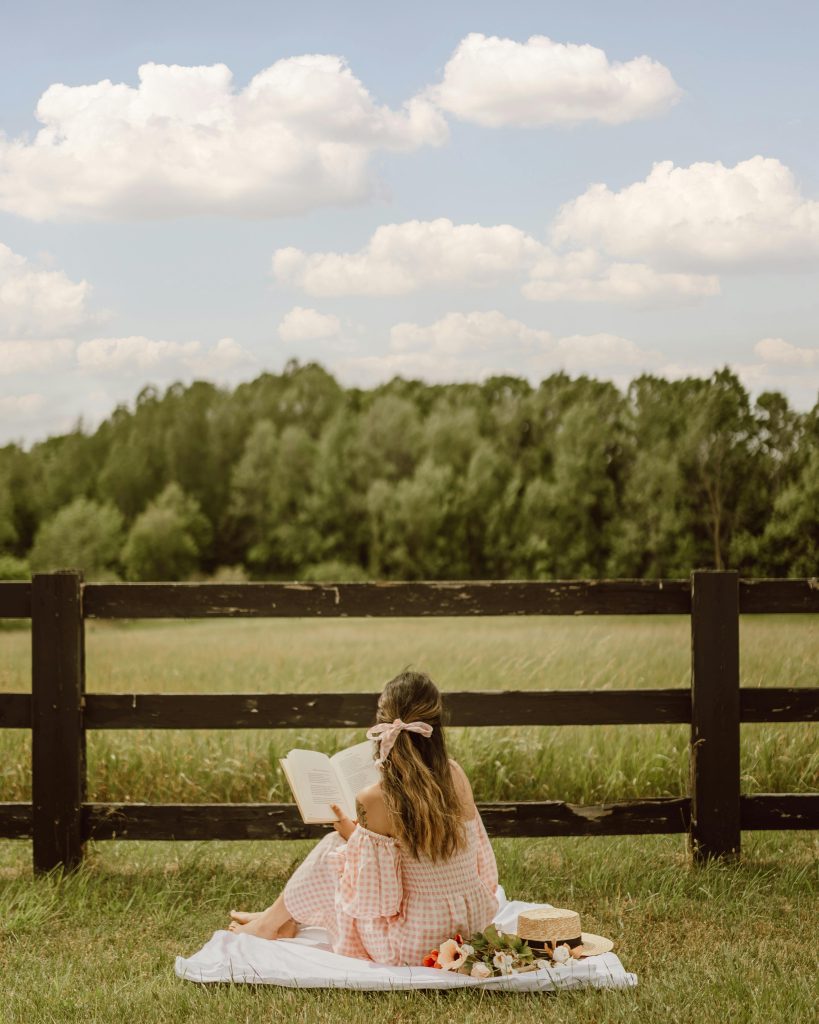 Woman reading on a picnic blanket by a wooden fence in the countryside.